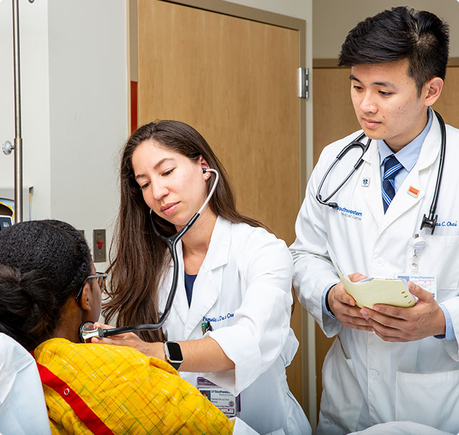 A doctor using a stethoscope on a patient in a hospital bed while another doctor takes notes